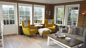 Una sala de estar acogedora con paredes de madera, ventanas amplias, dos sillones amarillos, un sofá gris y una mesa de centro con objetos decorativos.