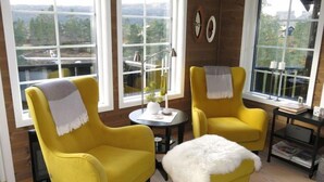 Una habitación con dos sillones amarillos, una pequeña mesa redonda y un otoman blanco y mullido. Hay dos ventanas con vistas a un bosque y un edificio.