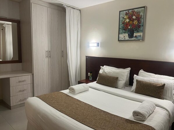 A neatly made bed with white linens and a brown throw, a wooden wardrobe, a nightstand with a lamp, and a painting of flowers on the wall.