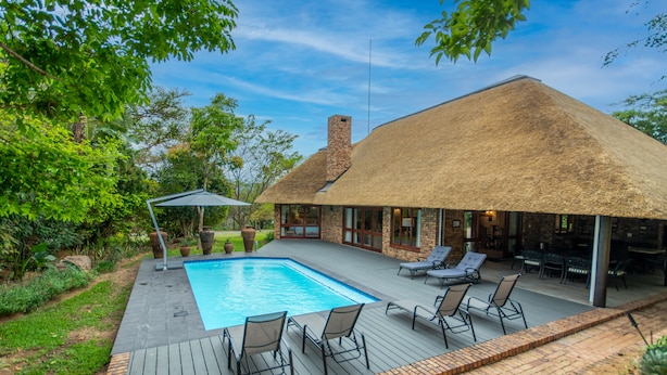 Ein Swimmingpool mit Liegestühlen, ein Gebäude mit Reetdach und eine Terrasse aus Ziegelsteinen.