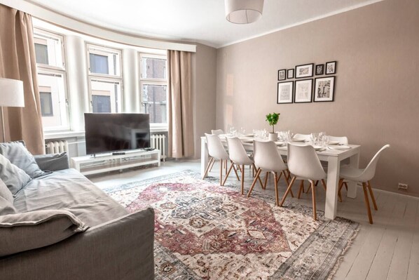 A modern living room with a sofa, TV, dining table with chairs, and a patterned rug. - 2ndhomes Bright & Spacious 5BR Apartment (Helsinki)