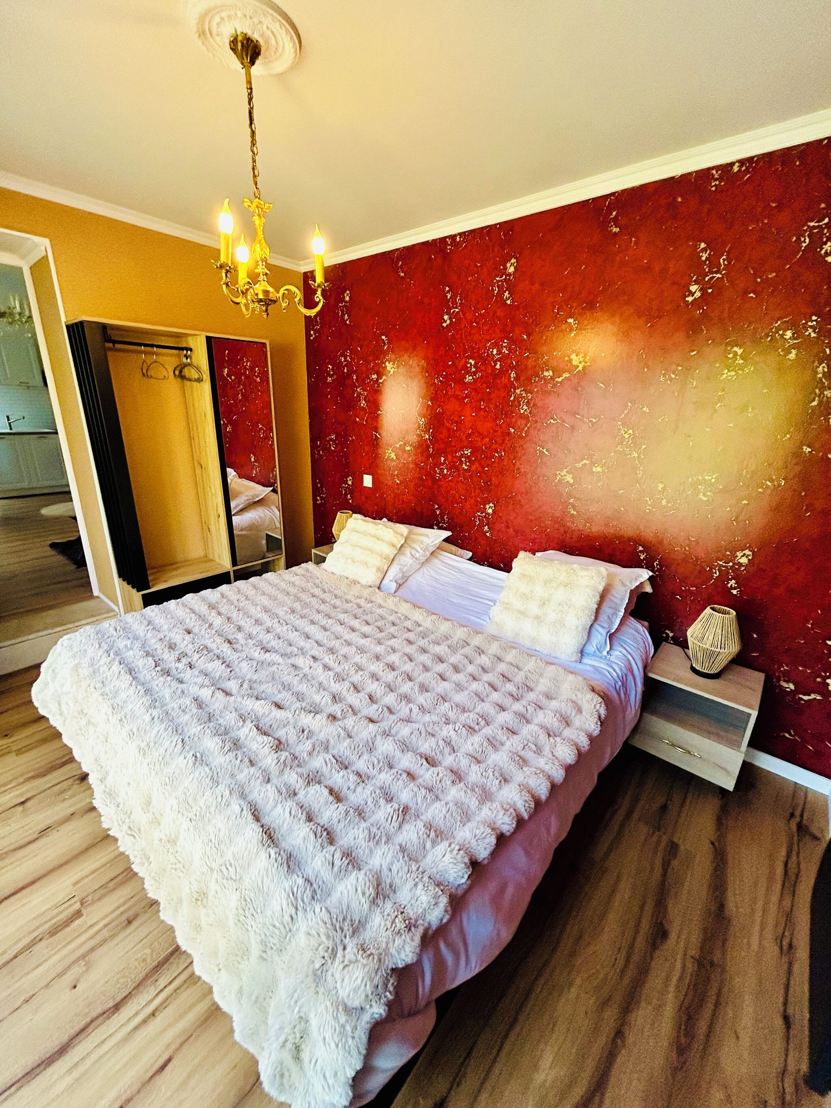 A bedroom with a large bed, a wooden floor, a red textured wall, and a chandelier.