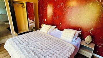 A bedroom with a large bed, a wooden floor, a red textured wall, and a chandelier.