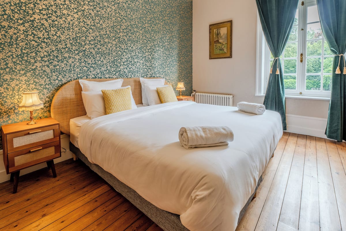 A neatly made bed with white linens, a wooden nightstand with a lamp, and a window with curtains.