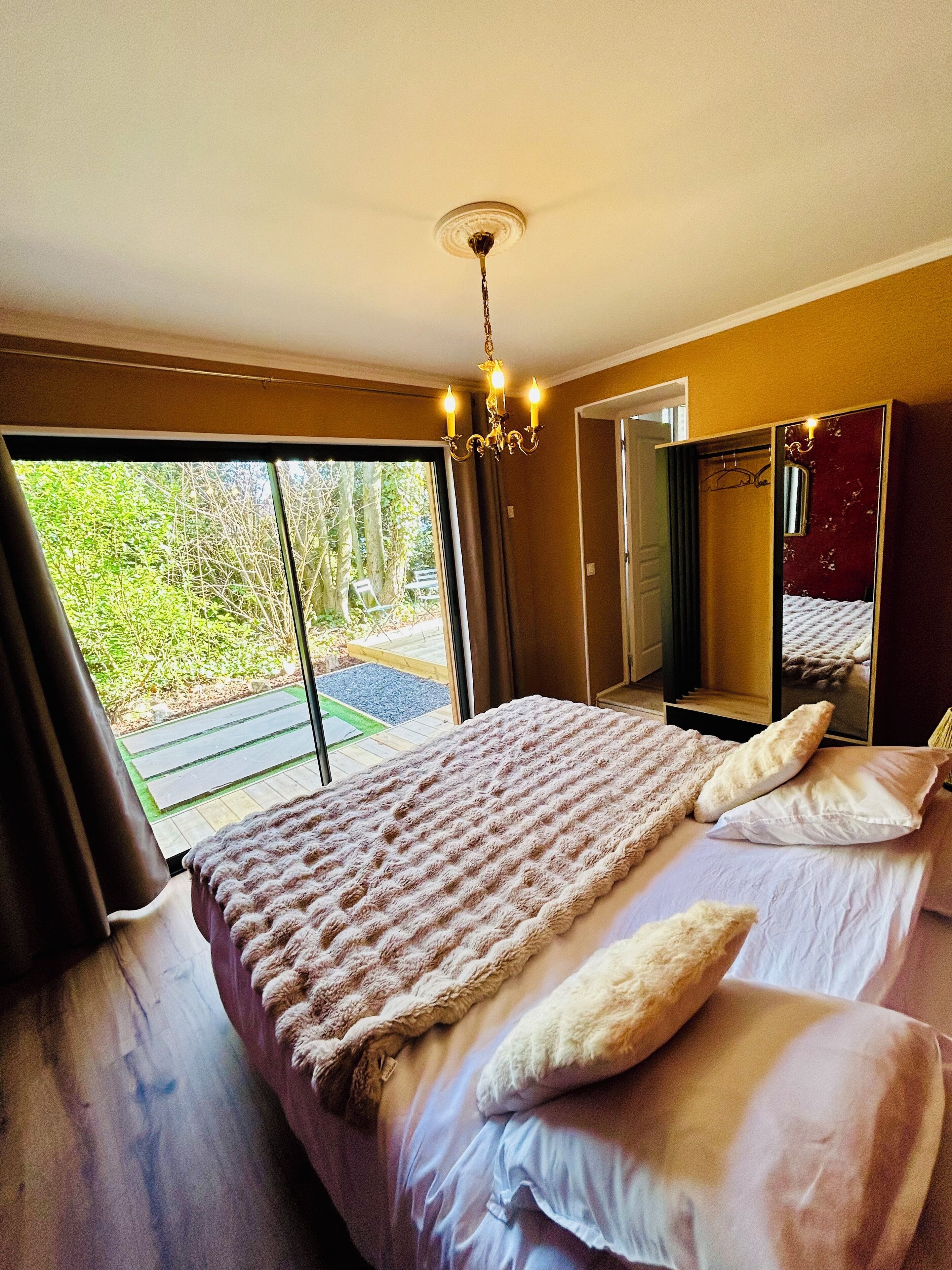 A bedroom with a large bed, a chandelier, a mirror, and a view of greenery outside.