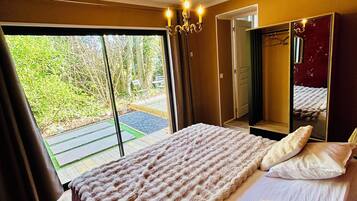 A bedroom with a large bed, a chandelier, a mirror, and a view of greenery outside.