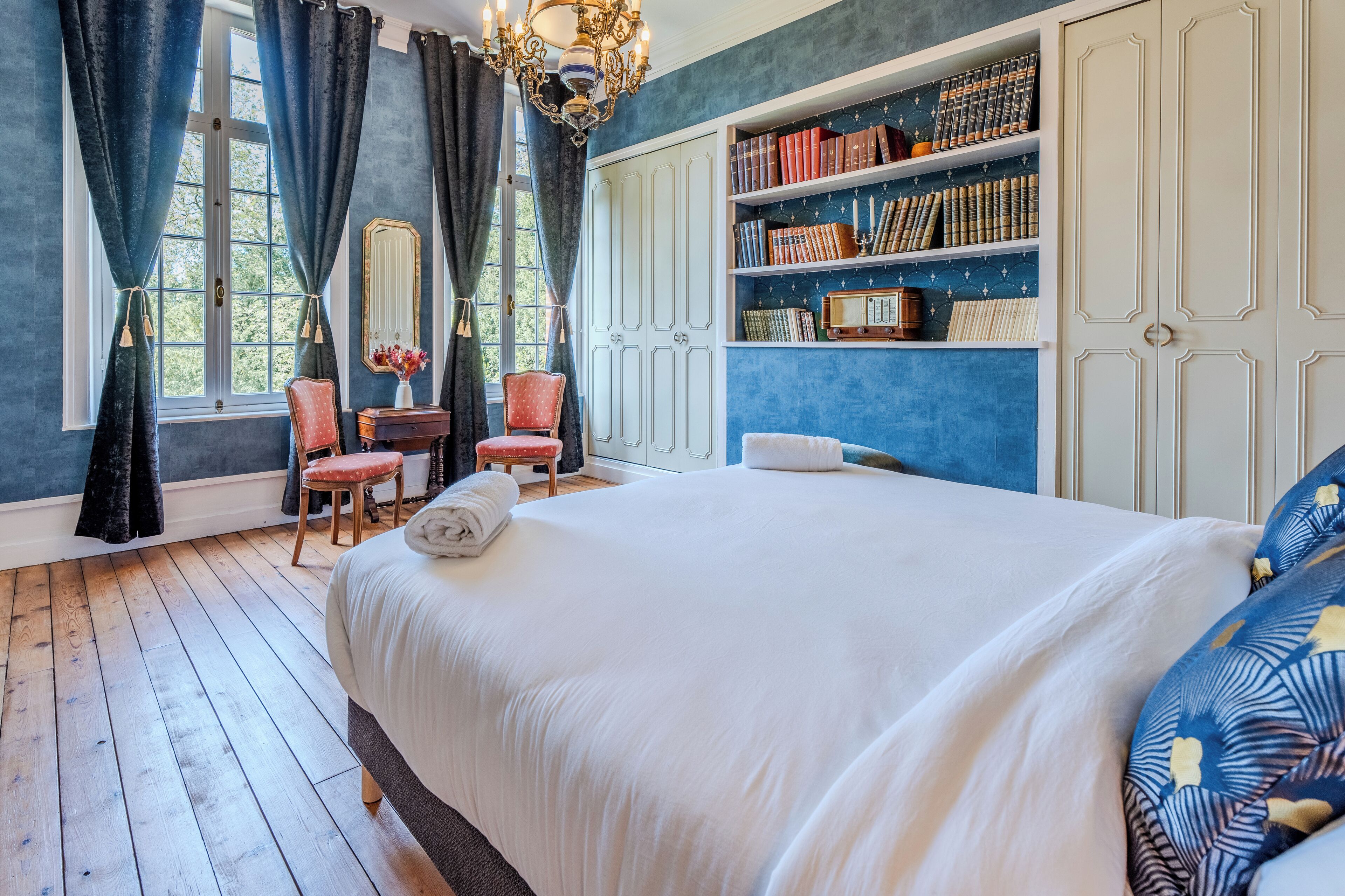 A bedroom with a large bed, a bookshelf, a chair, and a window with curtains.