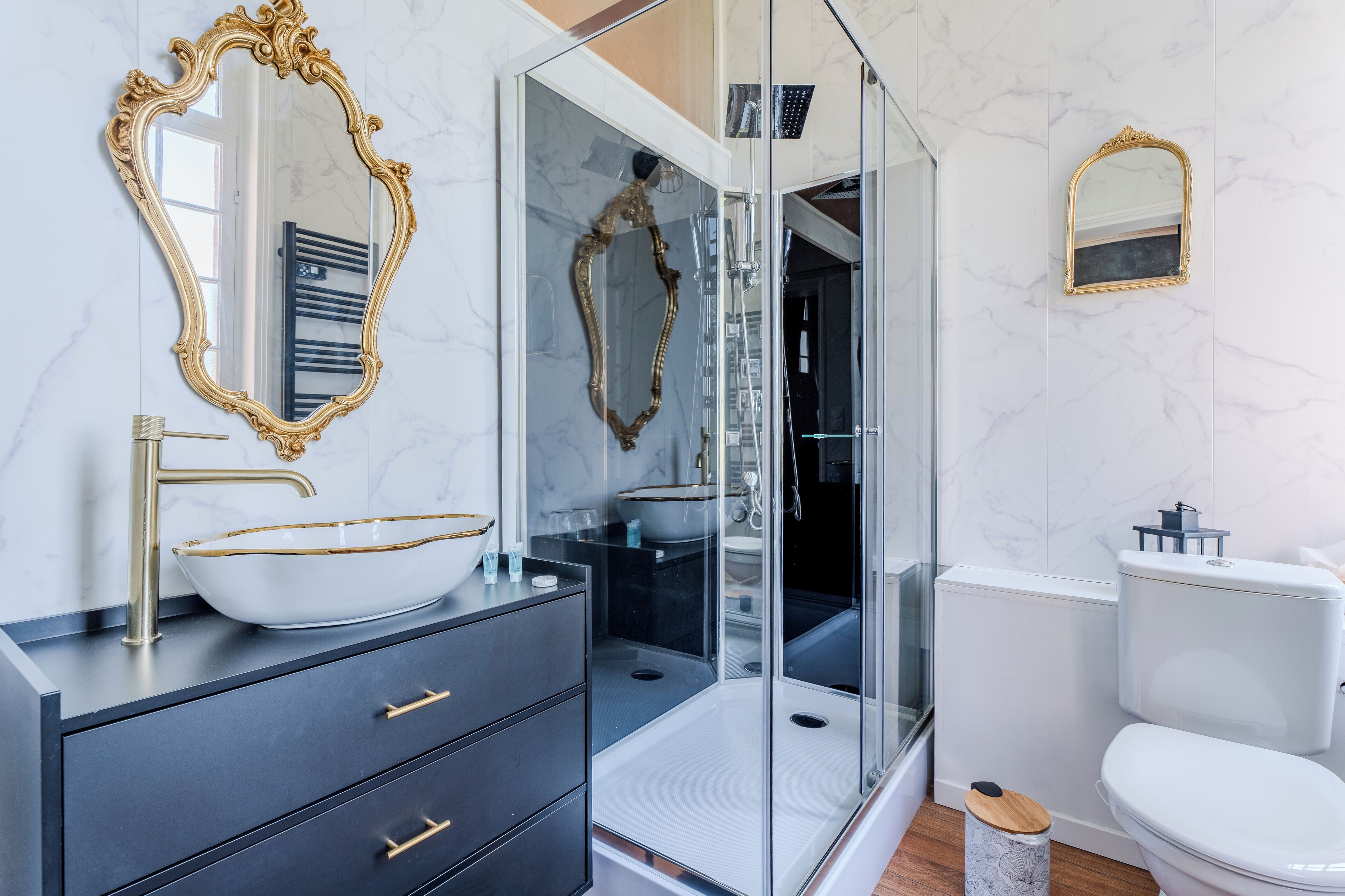 A modern bathroom with a glass-enclosed shower, a white toilet, and a vanity with a gold-framed mirror and a white vessel sink.