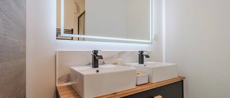 A modern bathroom with two white sinks, a mirror with integrated lighting, and a wooden vanity with a black cabinet.