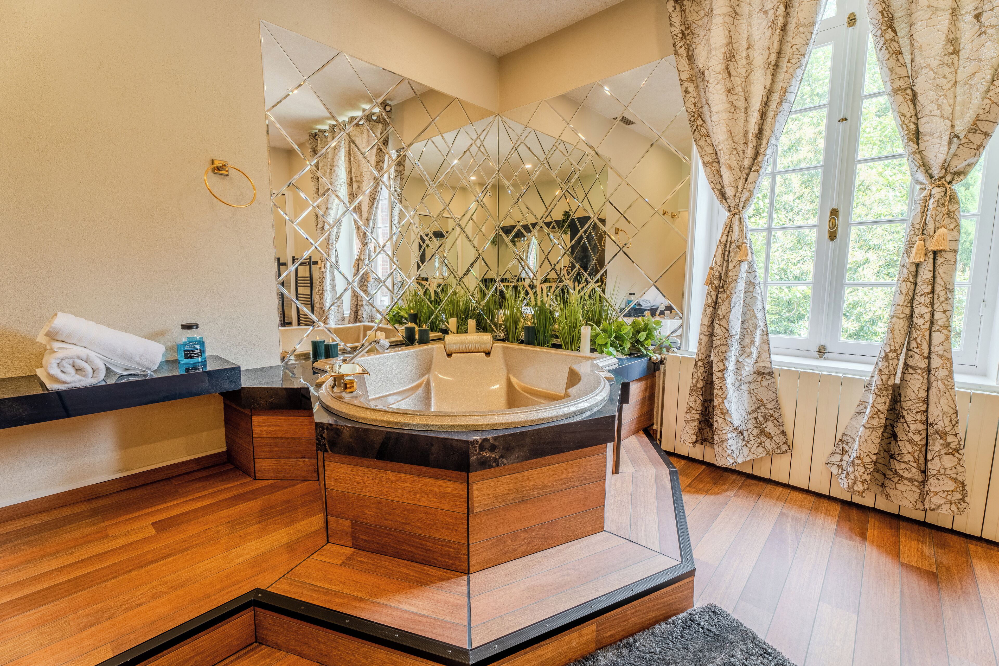 Une salle de bain moderne avec une baignoire sur pieds, un plancher en bois, un miroir à motifs géométriques et une fenêtre avec des rideaux.