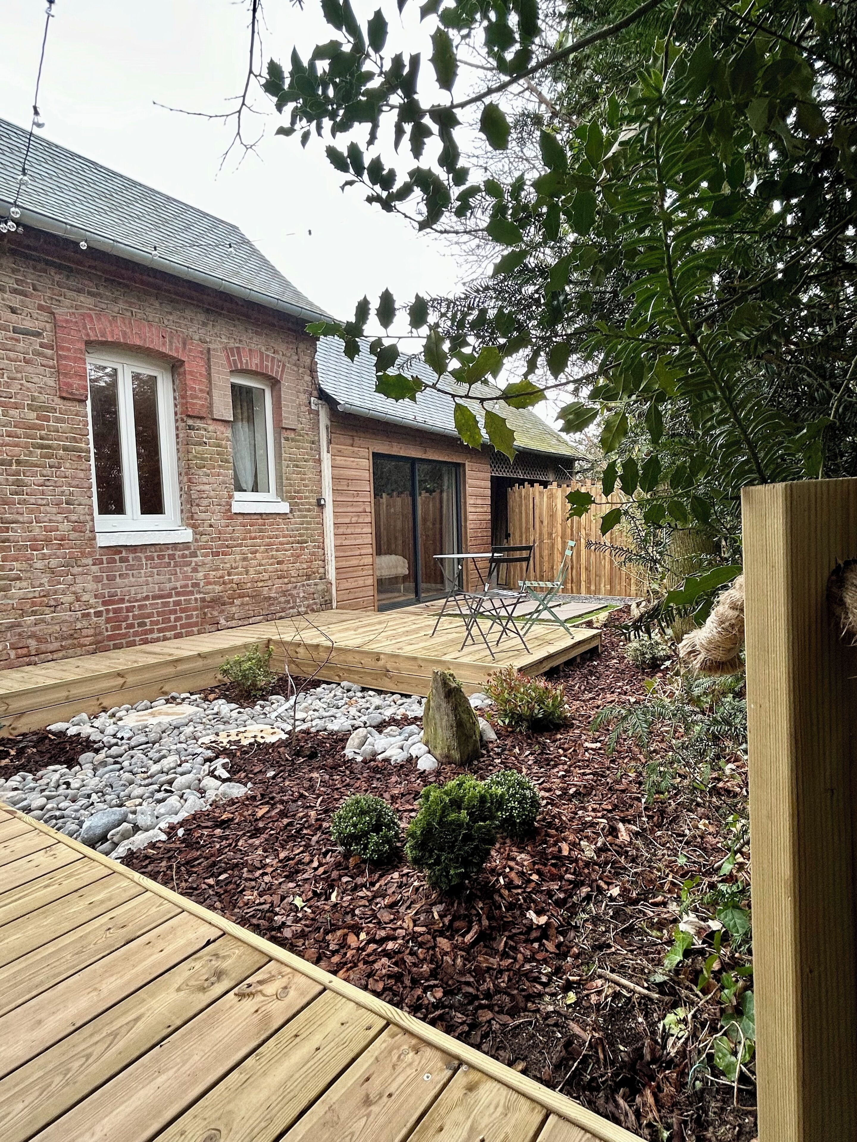 Une maison en briques avec une terrasse en bois, un petit jardin agrémenté de paillis et de rochers, et une clôture en bois.