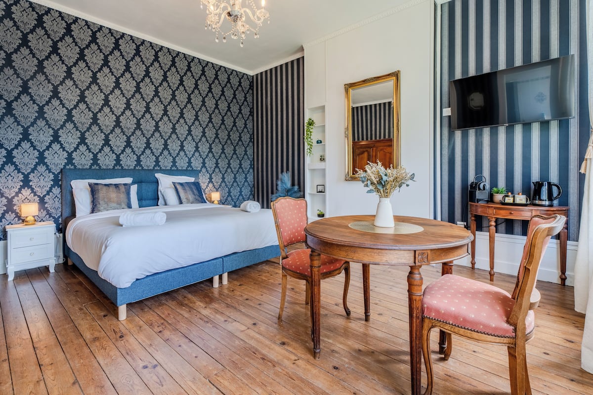 A bedroom with a bed, a round wooden table, two chairs, a nightstand, a TV, and a mirror.