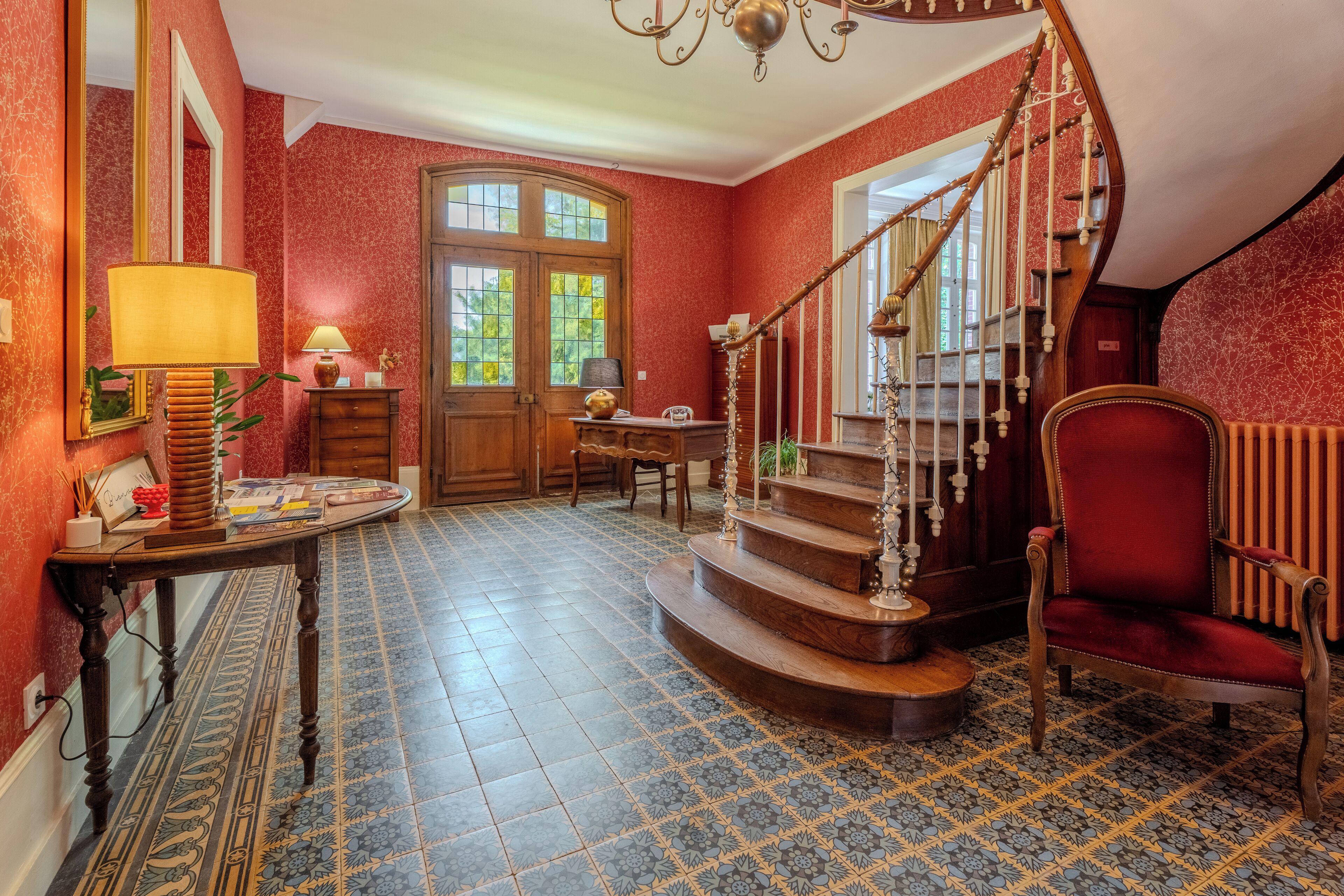 A room with a wooden staircase, a red chair, a table with a lamp, and a mirror on the wall.
