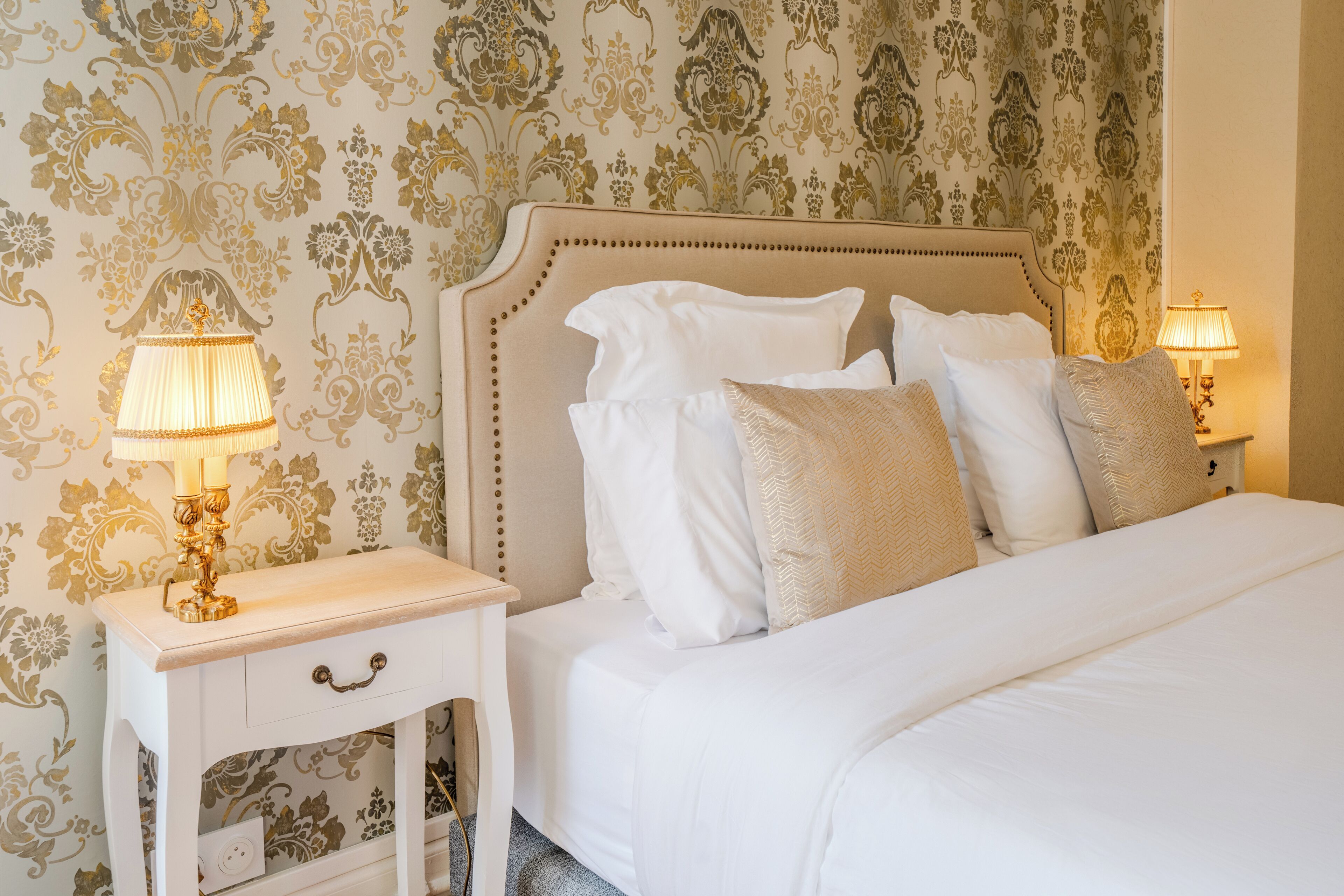 A neatly made bed with white linens and multiple pillows, a bedside table with a lamp, and a wall with ornate wallpaper.