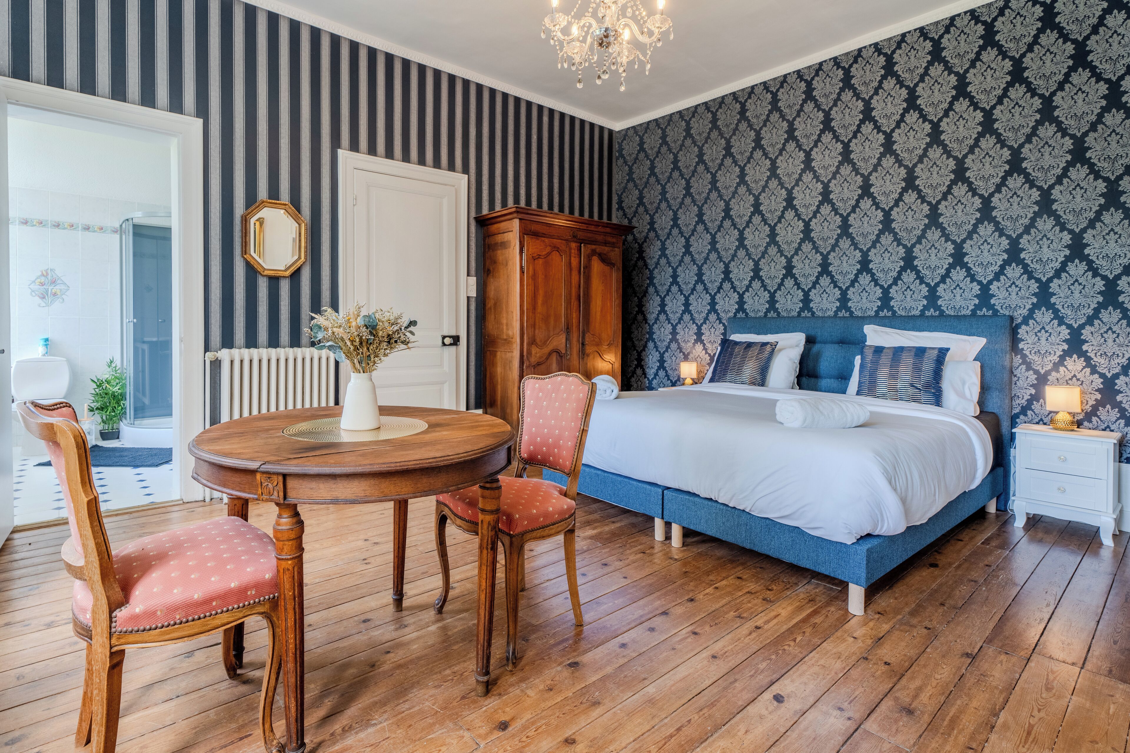 Un dormitorio con suelo de madera, cama con ropa de cama azul, armario de madera, mesa de comedor redonda con sillas y un espejo en la pared.