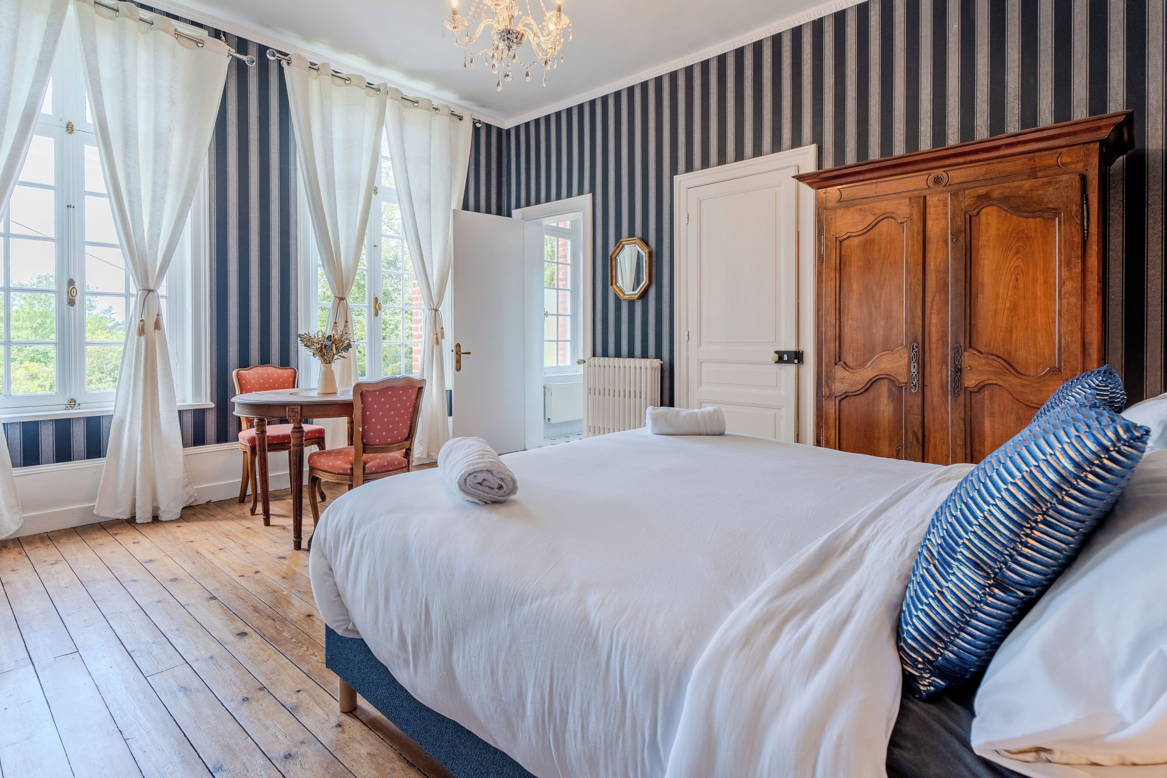 A hotel room with a large bed, a wooden wardrobe, a dining table with chairs, and a view through a window with curtains.