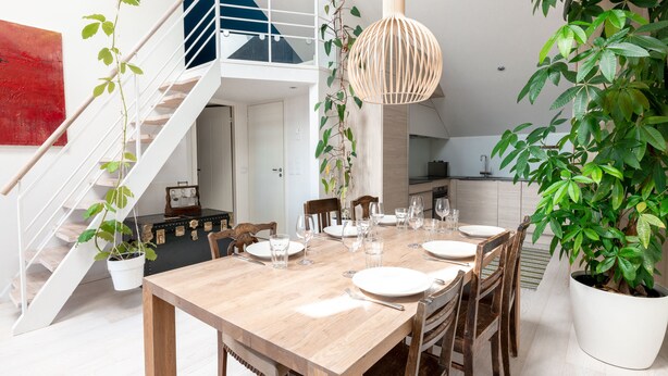 A dining area with a wooden table set for four, surrounded by chairs, a hanging light fixture, and potted plants. A staircase with a railing is visible in the background. - 2ndhomes 2BR Penthouse w Terrace&Sauna (Helsinki)