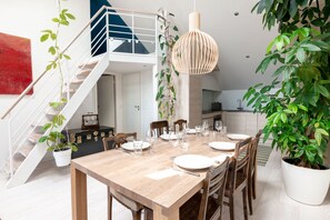 A dining area with a wooden table set for four, surrounded by chairs, a hanging light fixture, and potted plants. A staircase with a railing is visible in the background. - 2ndhomes 2BR Penthouse w Terrace&Sauna (Helsinki)