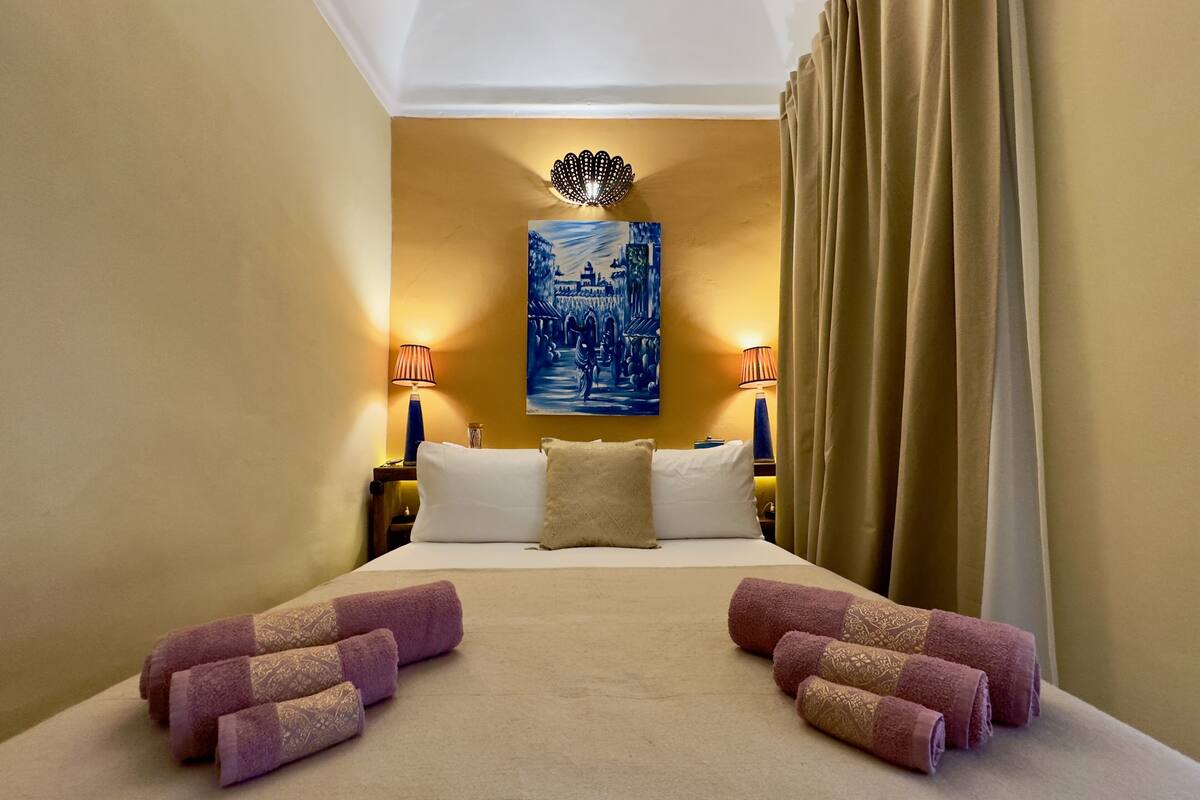 A hotel room with a large bed, two bedside lamps, a painting on the wall, and a window with curtains.