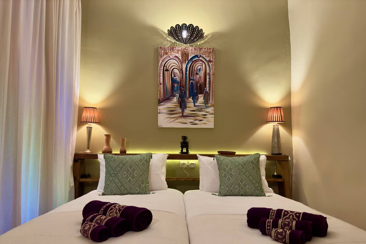 A hotel room with two beds, a painting of a street scene, two lamps, and a decorative light fixture.