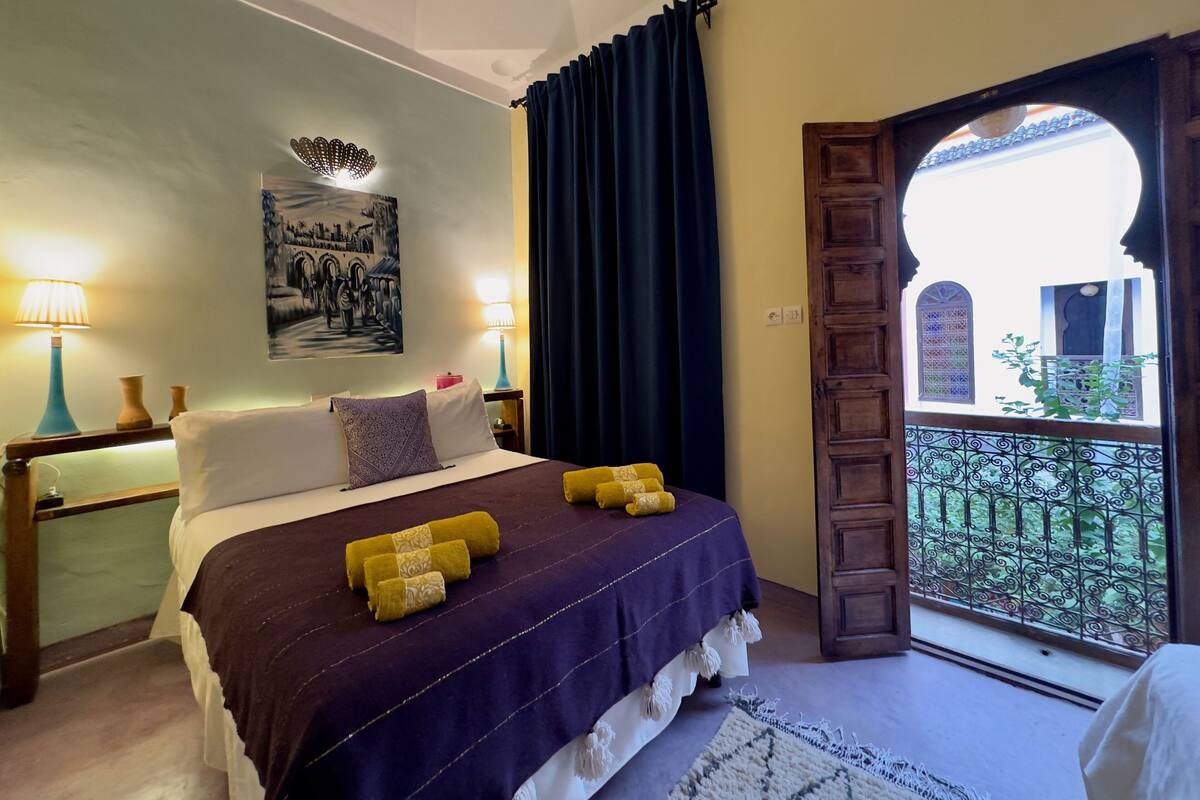 A bedroom with a bed, two bedside lamps, a painting, a window with a view, and a door leading outside.