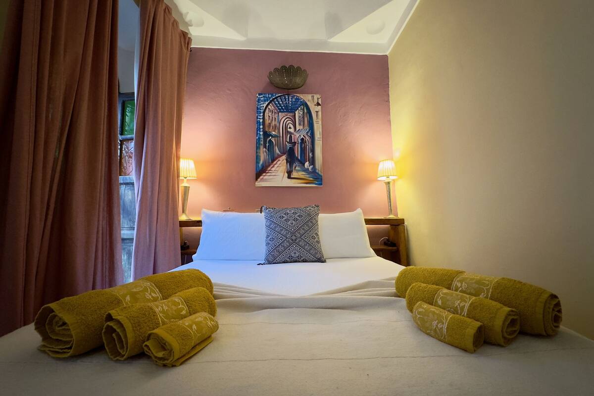 A hotel room with a bed, two lamps, a painting, and rolled towels.