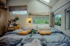 Un dormitorio con cama, almohadas, una mesita de noche con lámpara, una ventana con vistas y un tragaluz.