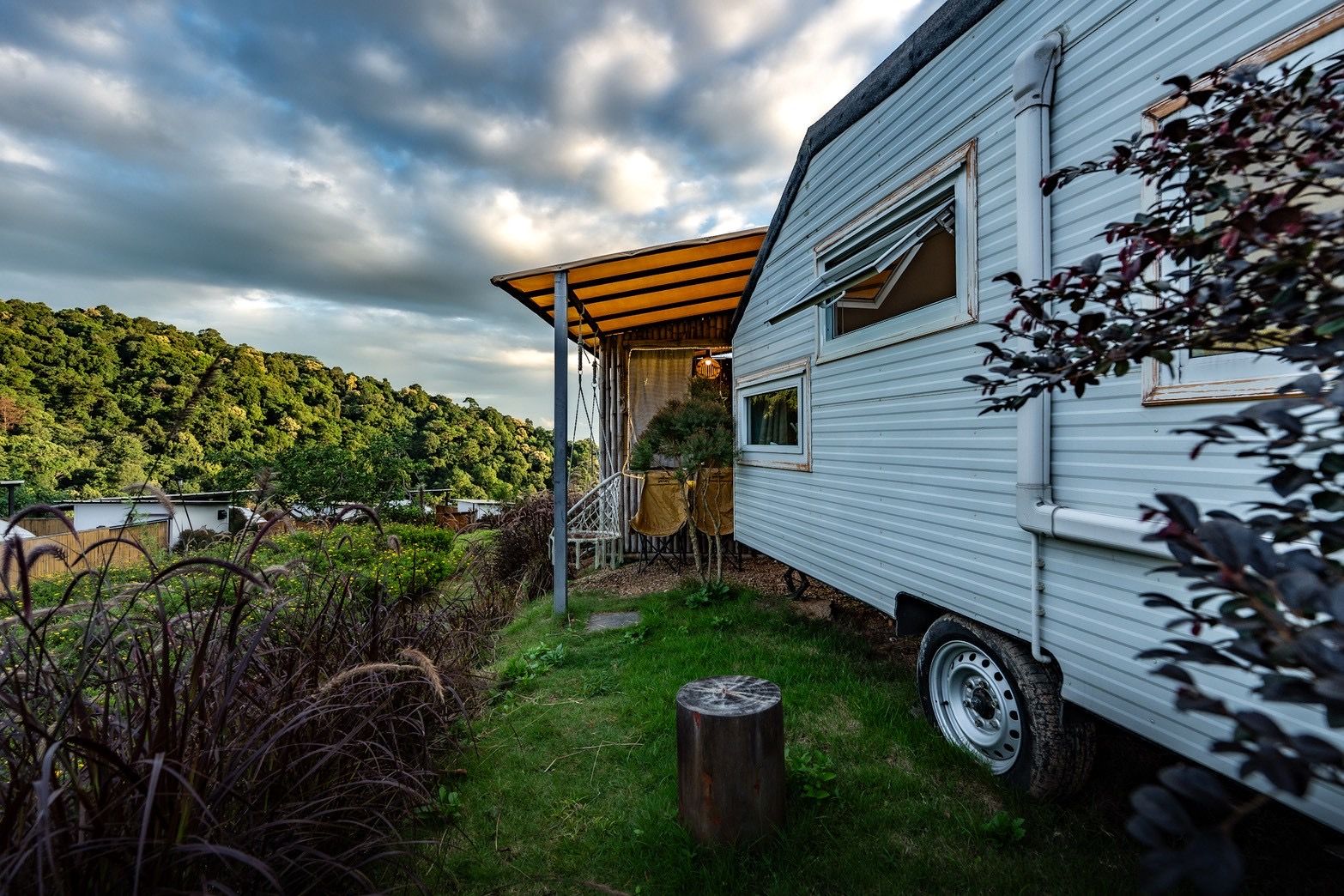 A mobile home with a porch, a small garden, and a view of a forested hill.
