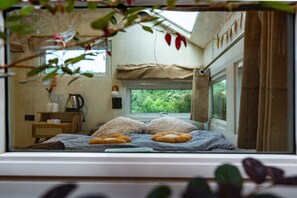 Un dormitorio con cama, almohadas, una ventana y vistas al paisaje verde exterior.