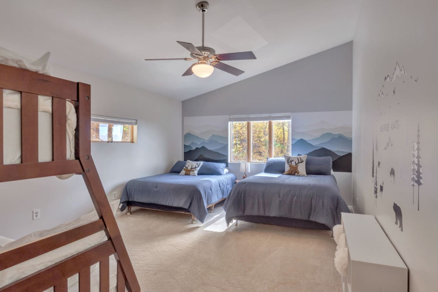 A bedroom with two beds, a ceiling fan, a window with a view of trees, and a wall with a mountain landscape mural.