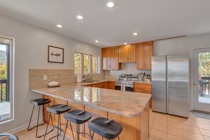 A modern kitchen with a central island, bar stools, wooden cabinets, stainless steel appliances, and a window with autumn foliage view.