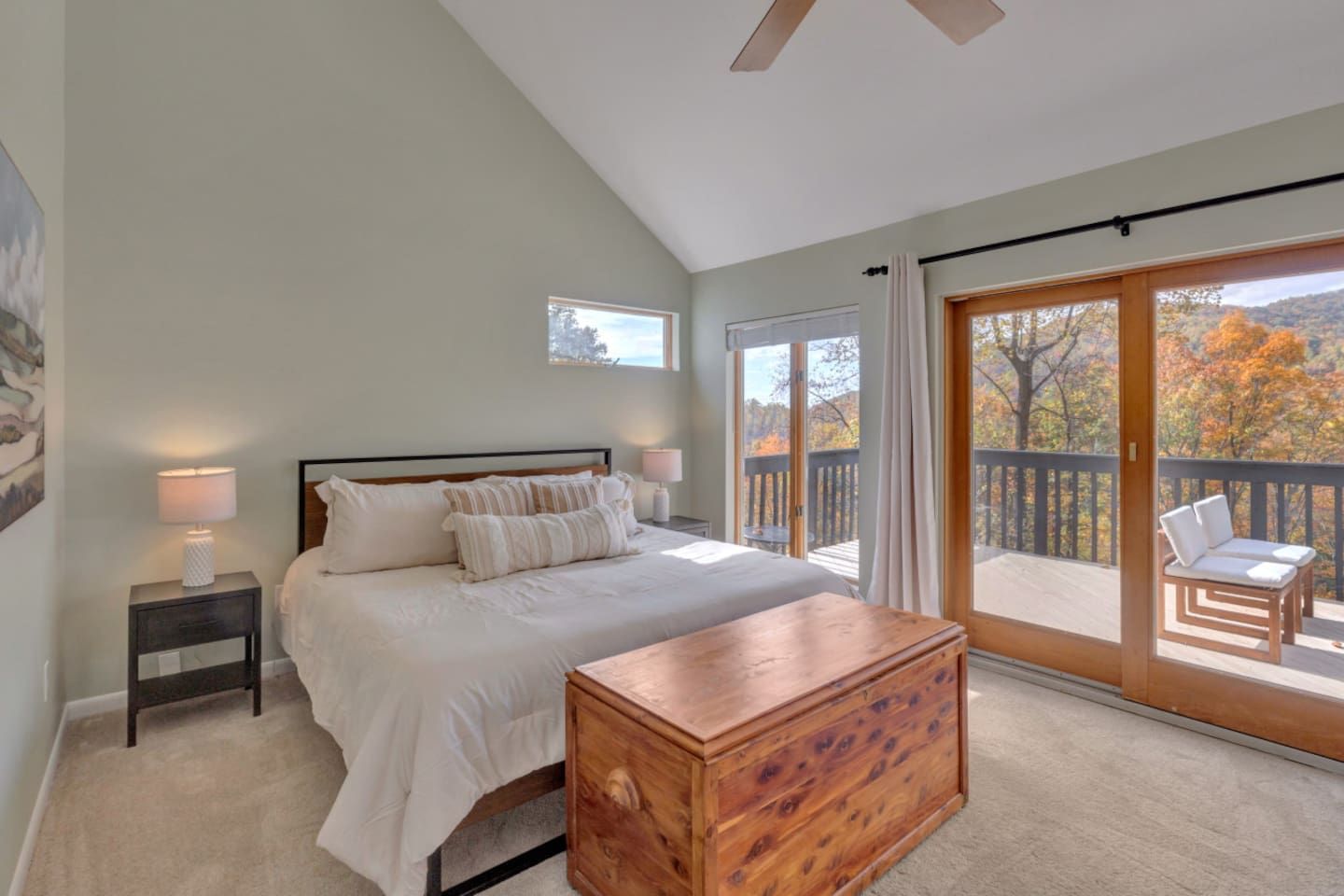 A bedroom with a bed, bedside tables, a wooden chest, a balcony with a chair, and a view of autumn trees.