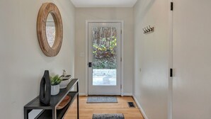 Een hal met een houten vloer, een witte deur met een geëtste ruit, een ronde spiegel, een zwart console tafeltje met een plant, en een klein tapijt.