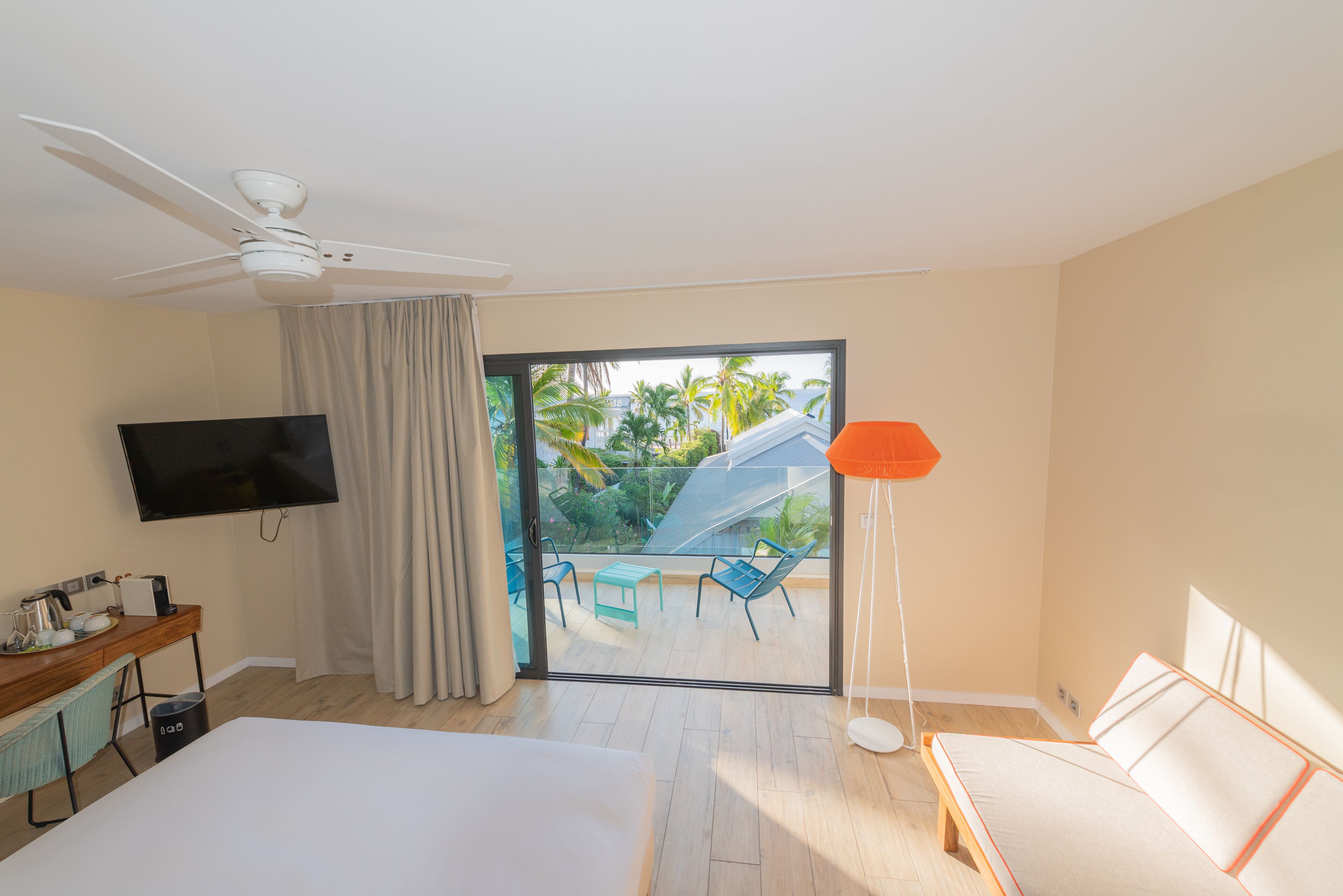 Ein Hotelzimmer mit einem Bett, einem Schreibtisch mit Flachbildfernseher, einem Deckenventilator, einer großen Schiebeglasdoor zu einem Balkon mit Sitzgelegenheit und einer Stehlampe.