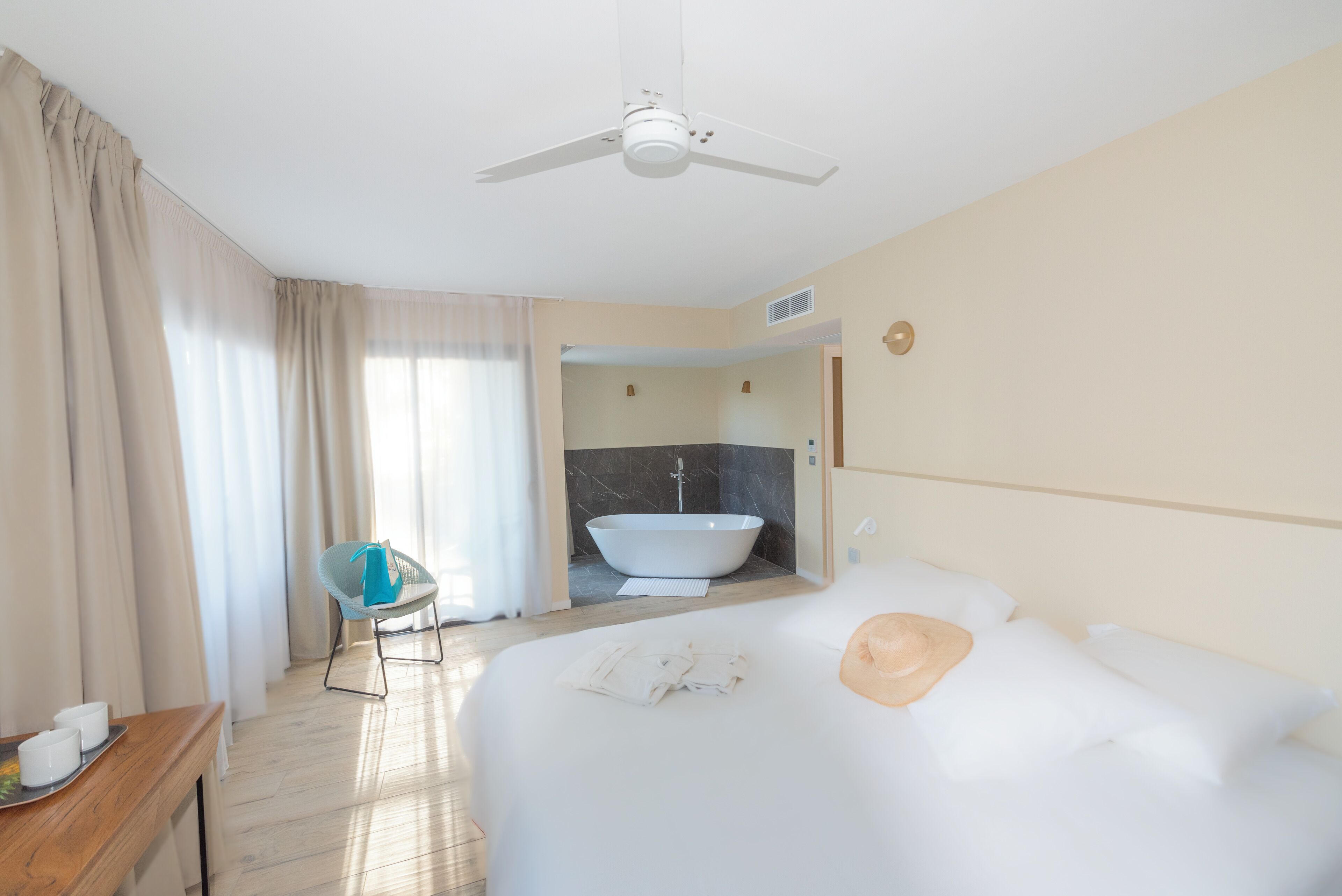 Une chambre d’hôtel avec un grand lit, un ventilateur de plafond, une salle de bain avec baignoire et un fauteuil.