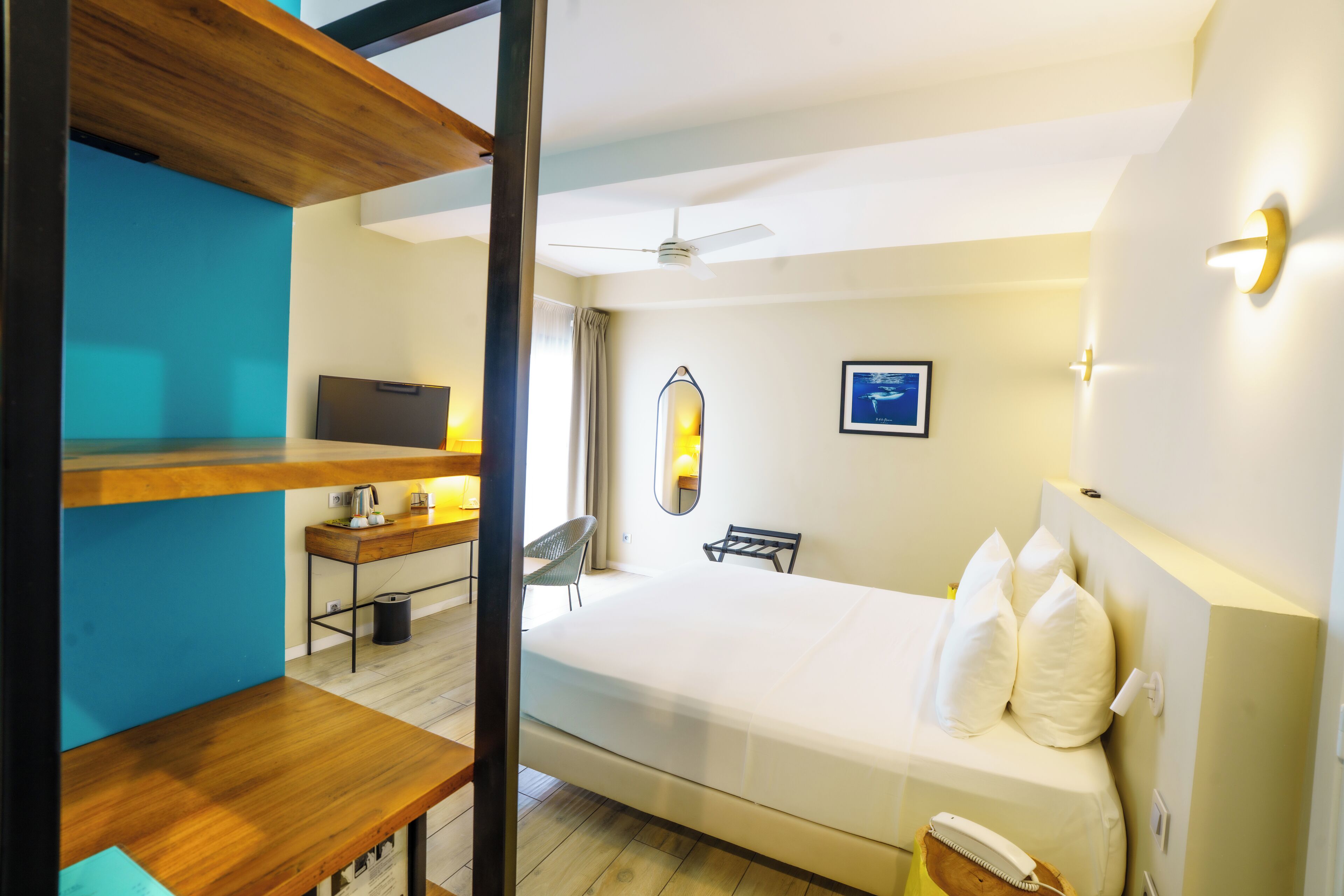 A hotel room with a bed, a desk with a computer, a chair, a mirror, and a ceiling fan.