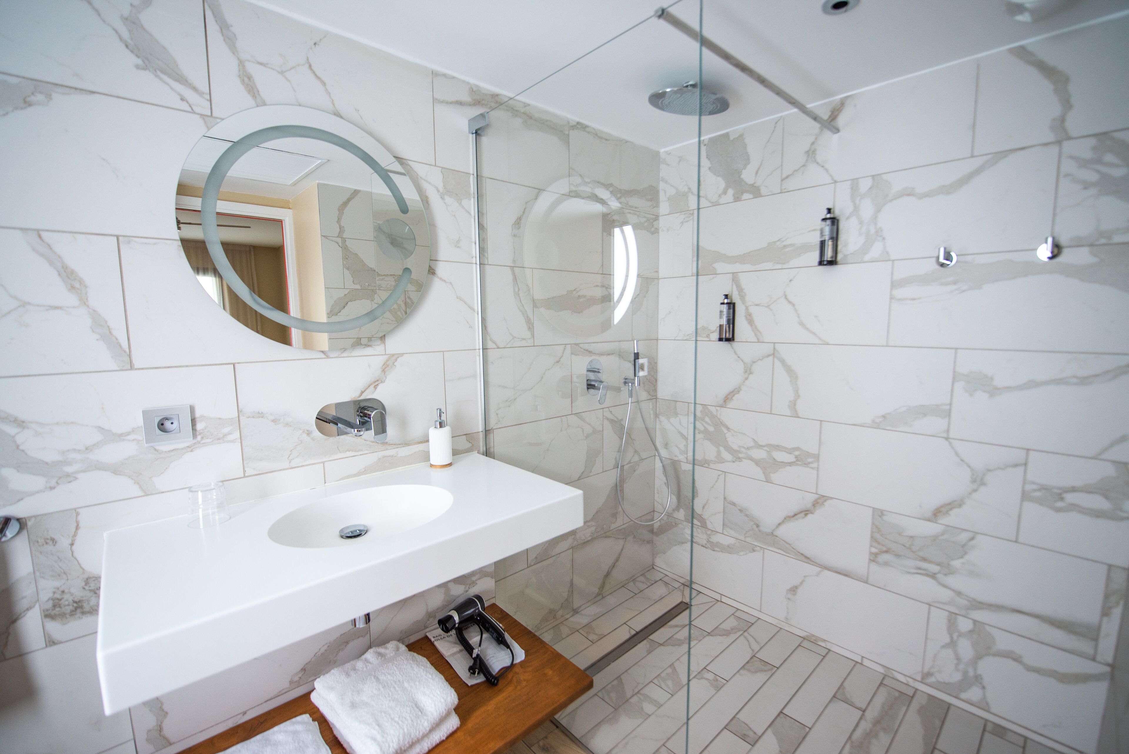 A modern bathroom with a marble-tiled wall, a white sink, a round mirror, and a glass-enclosed shower.