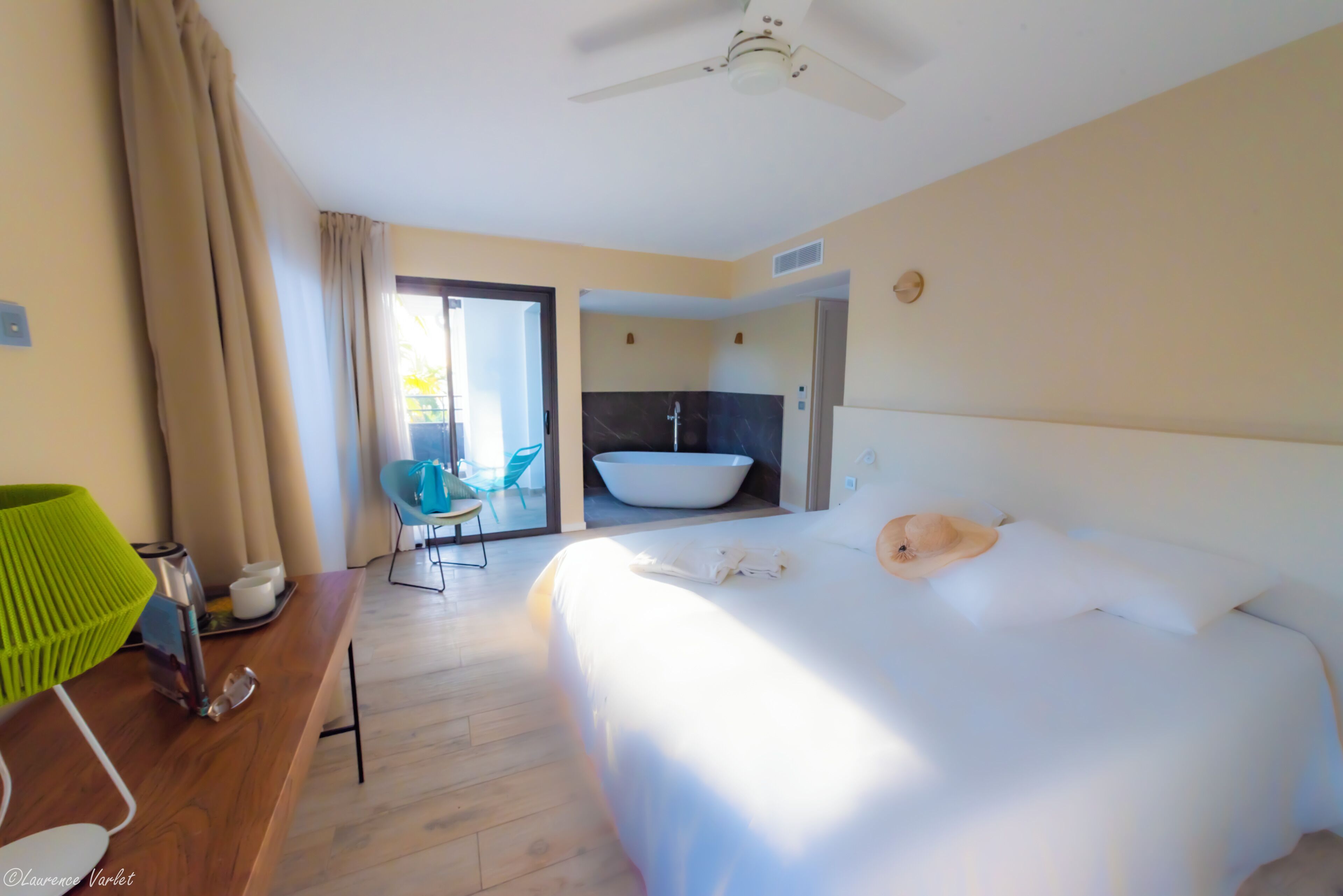 Una habitación de hotel con una cama, un escritorio con lámpara, una silla, un baño con bañera y un ventilador de techo.