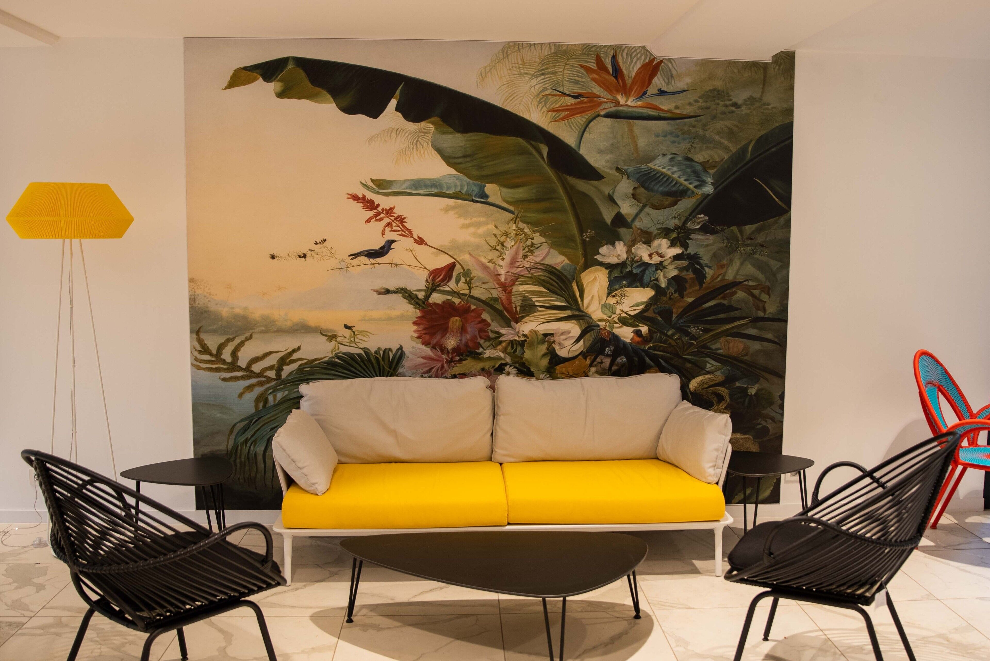 Una habitación con un sofá amarillo, sillas negras, un gran mural tropical y una lámpara de pie amarilla.