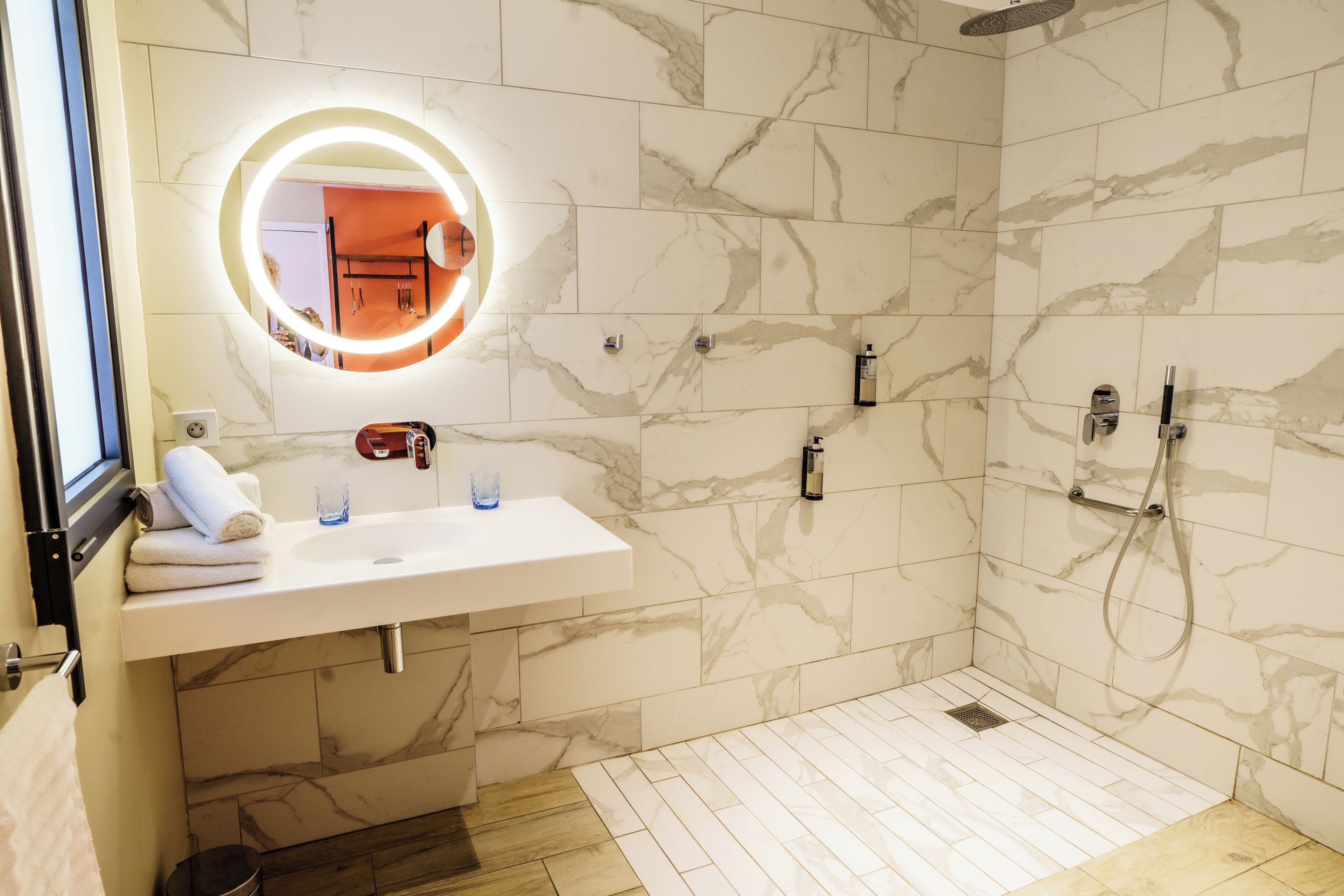 A bathroom with a marble-tiled shower, a white sink with a mirror, and a wooden floor.