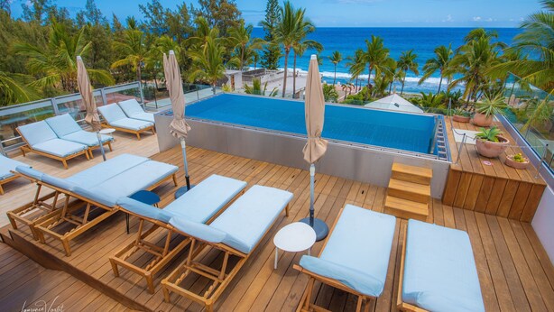 A rooftop pool area with lounge chairs and umbrellas, overlooking a beach and ocean. - Boutik Hotel 2A (Saint-Paul)