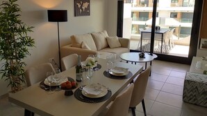 A dining area with a table set for two, a sofa, a potted plant, and a view of the outside balcony. - Luxe Appartement Orihuela-costa With Pool View (Orihuela)
