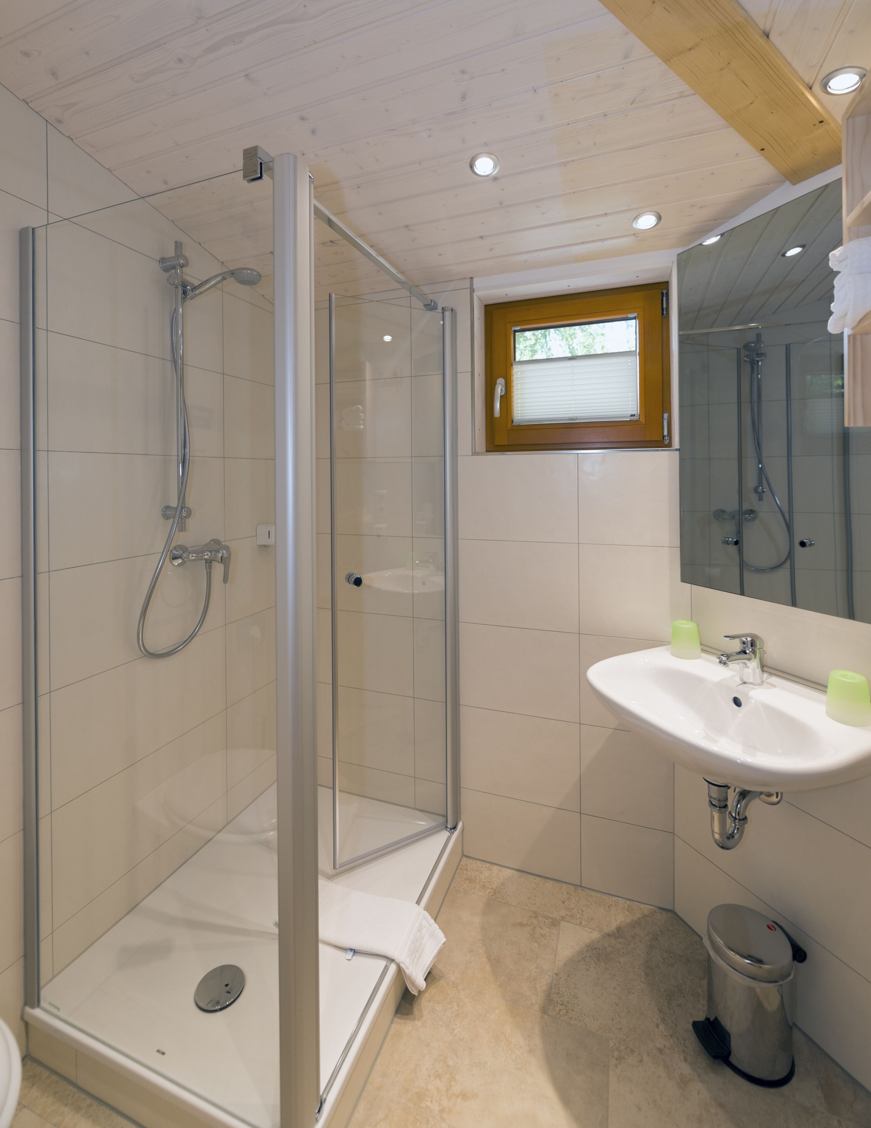 Een moderne badkamer met een glazen douchecabine, een witte wastafel met twee zeepdispensers en een klein raam met zonwering.