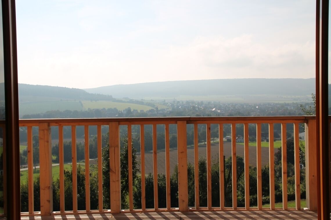 Ein Balkon mit Holzgeländer, der auf eine ländliche Landschaft mit Feldern und weit entfernten Hügeln blickt.
