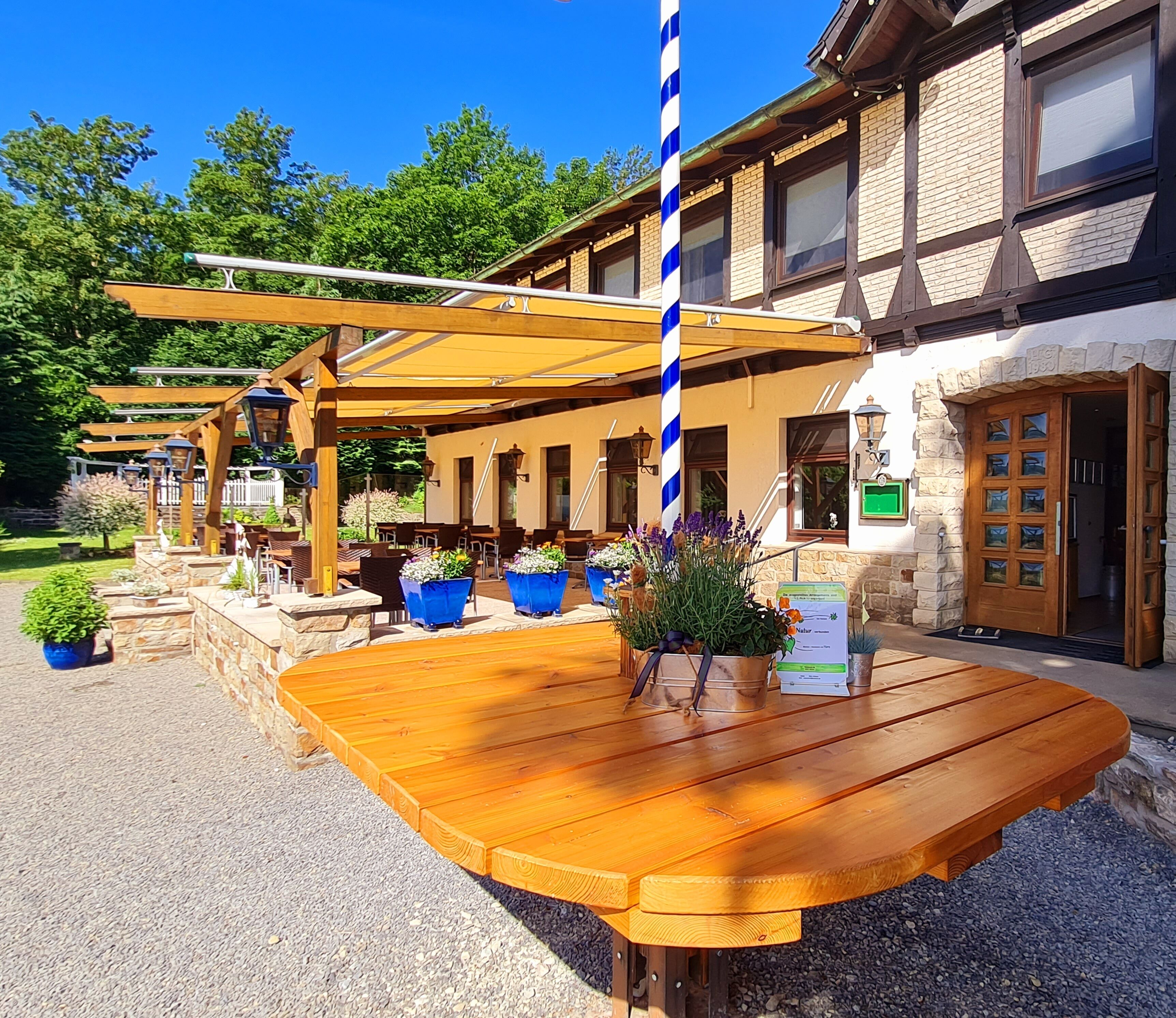 Un área de estar al aire libre con mesas y sillas de madera, rodeada de macetas con plantas y una pérgola.