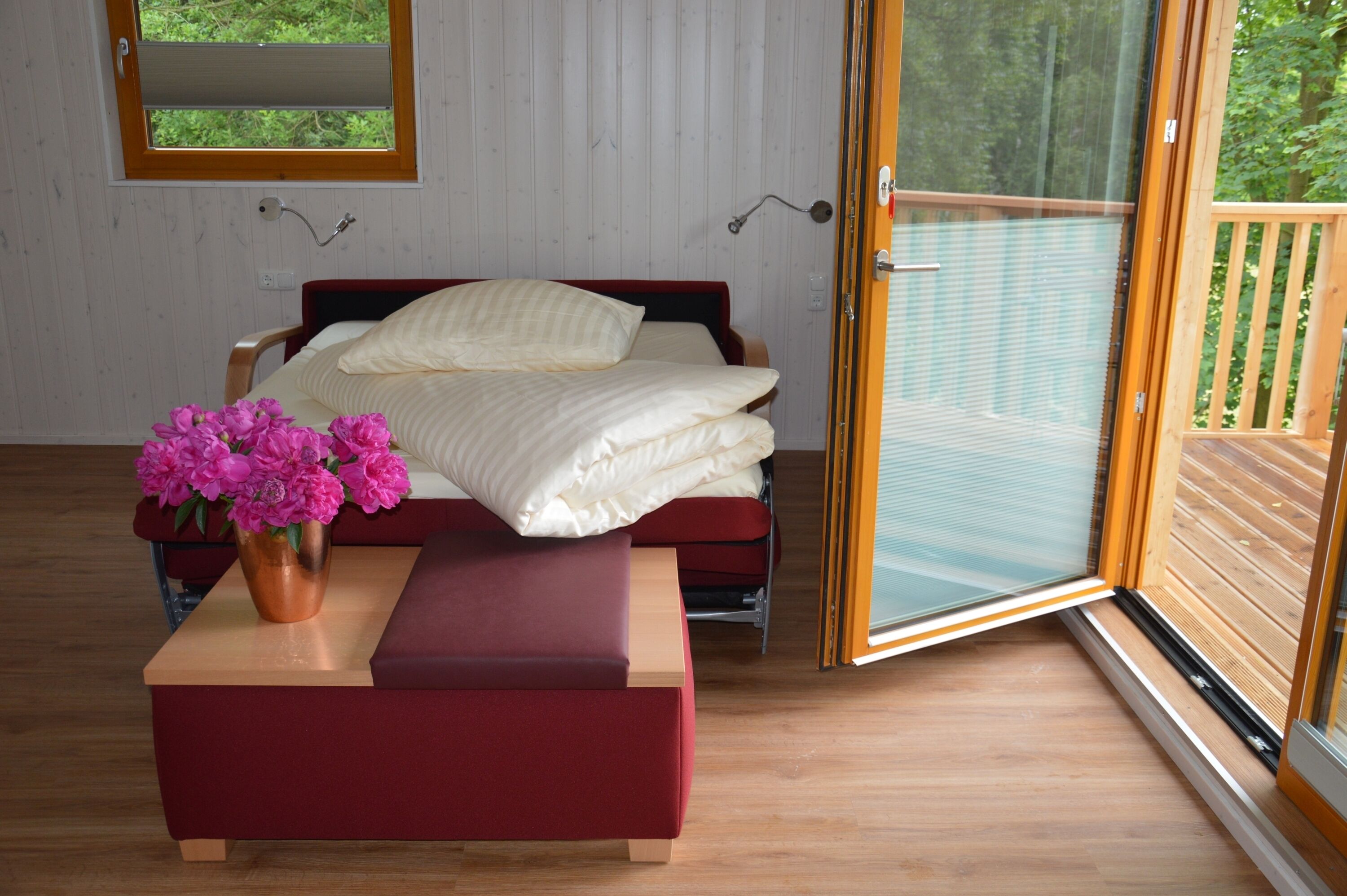 A bedroom with a bed, a wooden floor, a window, and a door leading to an outdoor deck.