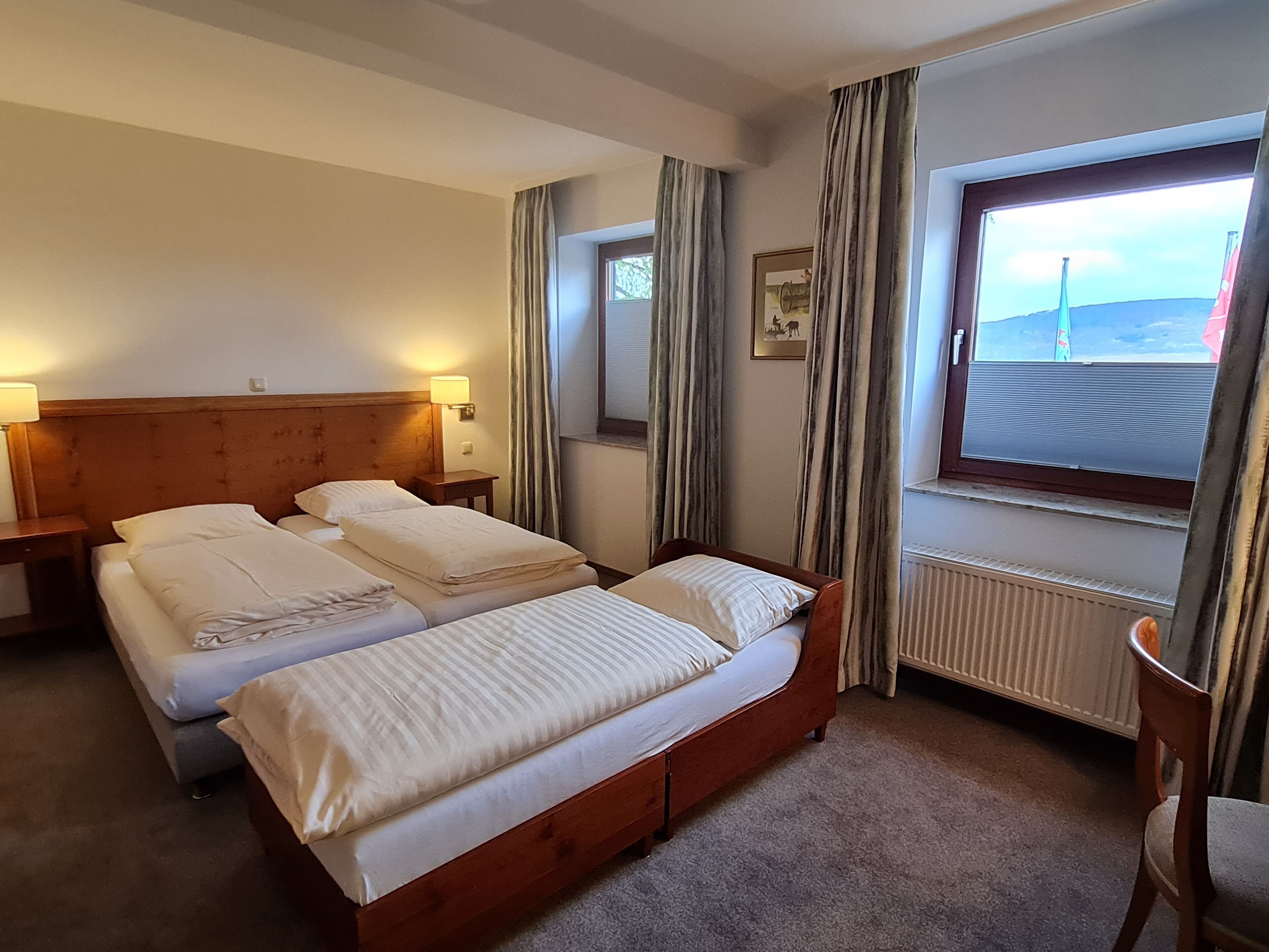 A hotel room with two beds, a nightstand with a lamp, a window with curtains, and a radiator.