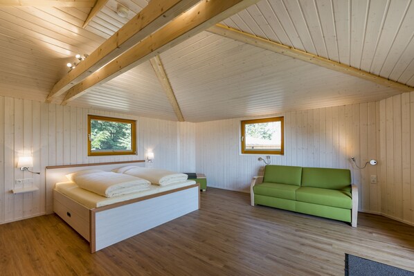 A wooden cabin interior with a bed, a green sofa, and two windows. - Hotel Waldquelle - Baumhaushotel Aerzen (Aerzen)