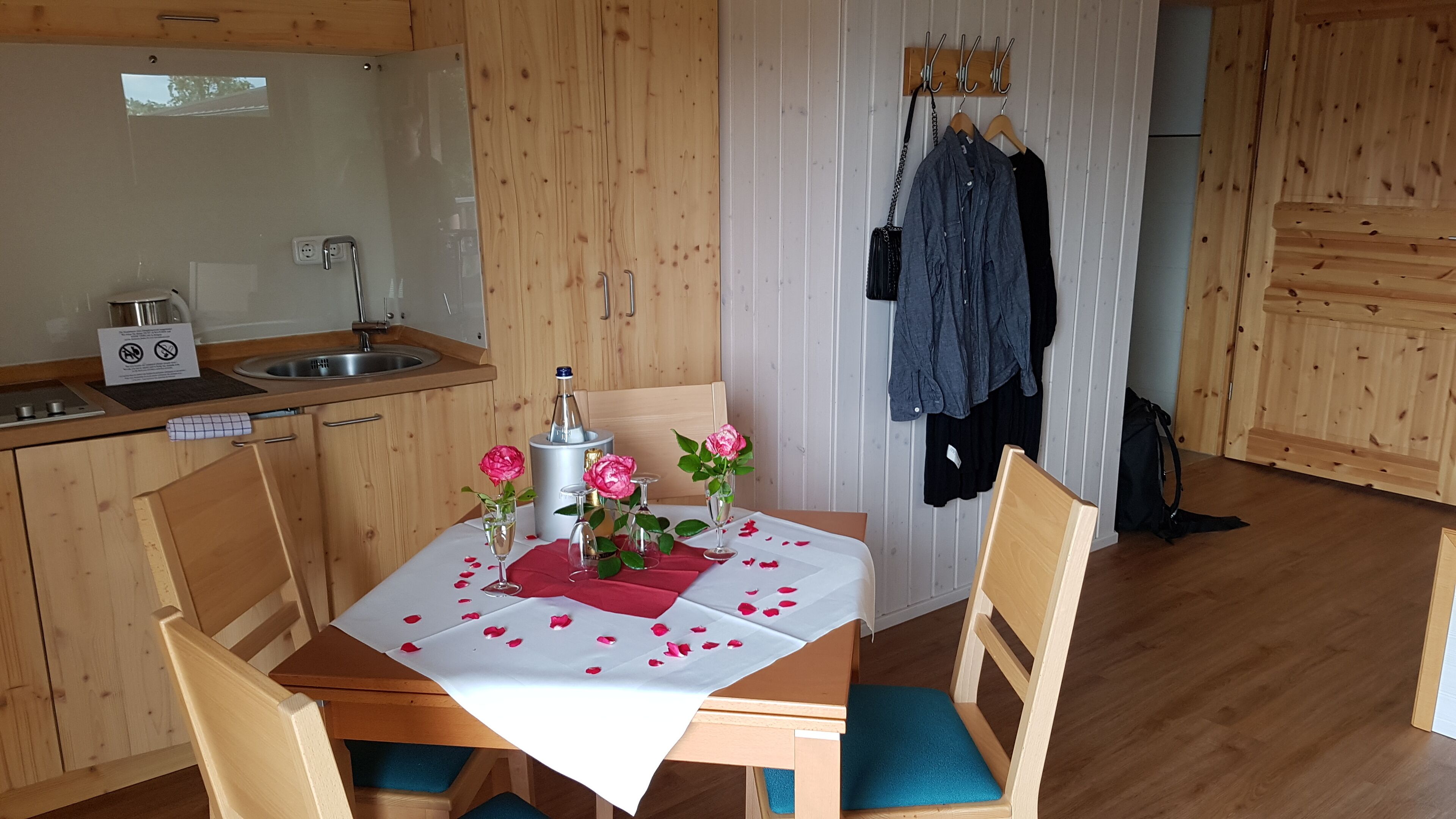 Una cocina pequeña con un lavamanos, una tetera y una mesa dispuesta para dos, decorada con pétalos de rosa y una botella de champán.