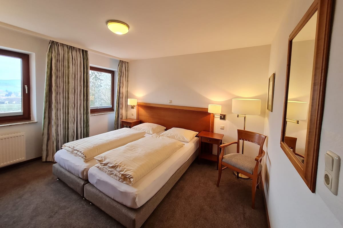 Ein Hotelzimmer mit zwei Einzelbetten, einem Holzkopfende, Nachttischlampen, einem Sessel, einem großen Spiegel und einem Fenster mit Vorhängen.